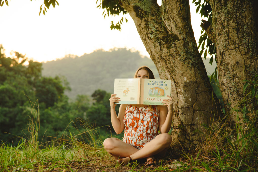 Mulher grávida sentada ao pé de uma árvore, segurando um livro infantil que lê para o bebê.